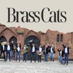 Die Band BrassCats steht vor einer Kirche mit ihren Instrumenten und springt dabei in die Luft